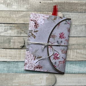 Op de afbeelding: Een decoratieve, gevouwen papieren envelop met een bloemen- en hertenpatroon. De envelop is vastgebonden met zilverkleurig touw en heeft een rood lint. Het ontwerp toont rozen, poinsettia's en herten op een grijze achtergrond.