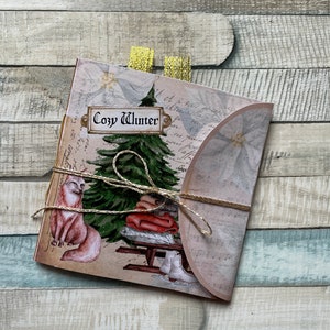 Op de afbeelding: Een gevouwen kaart met een winterthema. De kaart toont een aquarel illustratie van een vos, een kerstboom en een stapel dekens op een slee. De tekst "Cozy Winter" staat op de kaart. De kaart is vastgebonden met touw en heeft een gouden lint.