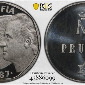 May include: A silver-toned commemorative coin showing the profiles of Juan Carlos and Sofia on the obverse with the text JUAN CARLOS I Y SOFIA 1987. The reverse features a crown above the letter M, the word PRUEBA, and a star with the number 87.