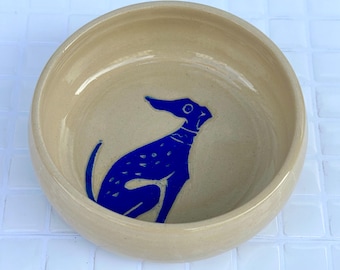 May include: A round, beige ceramic bowl with a smooth, glossy finish. The interior base of the bowl features a bold, dark blue graphic illustration of a sitting dog with a spotted pattern on its body and a collar around its neck.