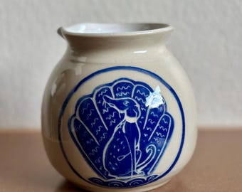 May include: A small, cream-colored ceramic vase with a blue illustration of a rabbit inside a stylized shell. The vase has a rounded body and a slightly flared rim. The rabbit is depicted in a seated position.