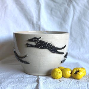 Könnte beinhalten: Eine cremefarbene Keramikschale mit einem schwarzen Hunde-Design. Die Schale hat eine weite Öffnung und einen abgerundeten Boden. Mehrere gelbe Früchte liegen neben der Schale auf einem weißen Tuch.