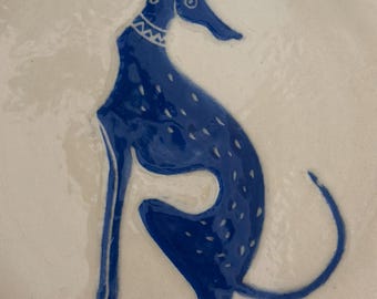 May include: A ceramic plate featuring a blue illustration of a dog. The dog is depicted in a seated position with a long neck and tail. The plate has a white background with a blue rim.