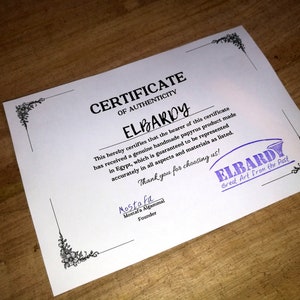 May include: A white certificate of authenticity with black text and a purple stamp. The certificate is for a handmade papyrus product from Elbardy, "Great Art from the Past".