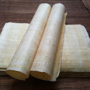 May include: Two rolls of natural beige papyrus paper, unfurled and stacked on a wooden surface.