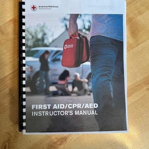 May include: A spiral-bound manual with the text "FIRST AID/CPR/AED INSTRUCTOR'S MANUAL" and the American Red Cross logo. The cover features a person holding a red first aid kit, with a blurred background of people and a vehicle.