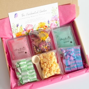 May include: A cardboard box lined with pink tissue paper, filled with packets of colourful ingredients and a wooden spoon. The box includes a card that reads "The Enchanted Garden" with floral illustrations. Packets contain green, pink, and blue items.
