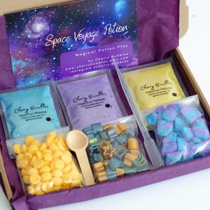 May include: A boxed kit labeled "Space Voyage Potion" with various colorful components. Includes packets of blue, purple, and yellow powders, plus star-shaped and other shaped soaps, a wooden spoon, and a dropper. The box has a purple interior.