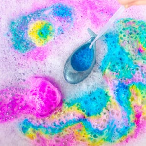 May include: A bath bomb dissolving in water, creating colorful swirls of pink, blue, yellow, and green. A silver-colored, whale-shaped bath product is held above the water, filled with blue foam. Bubbles and foam surround the bath bomb.
