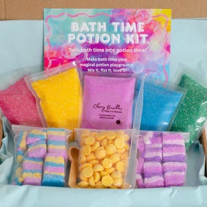 May include: A bath time potion kit with colorful ingredients in clear bags. The kit includes pink, yellow, blue, and green granules, along with yellow disc-shaped items and striped bath bombs. A sign reads "BATH TIME POTION KIT".