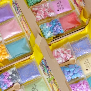 May include: A box filled with small, clear bags containing colorful bath bomb ingredients. The bags hold various shapes and colors, including pink, blue, purple, and yellow. Small wooden scoops are included. The box is lined with yellow paper.