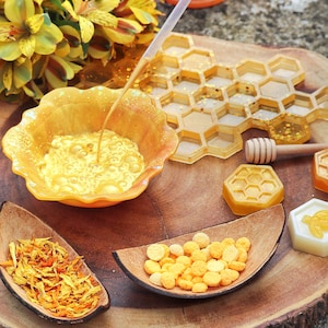 May include: A wooden tray with a honeycomb mould, a small bowl of yellow liquid, a bowl of yellow flower petals, a bowl of yellow round sweets, and a wooden honey dipper.