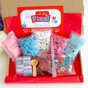 May include: A red and white potion kit with the text "Red white and Fizz! Potion Kit". The kit includes various ingredients in clear bags, such as pink and blue sprinkles, star-shaped candies, and bath bombs. A wooden spoon and dropper are also included.