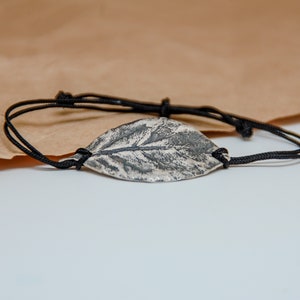 May include: A silver leaf charm bracelet with a black cord. The leaf is textured and has a detailed vein pattern.