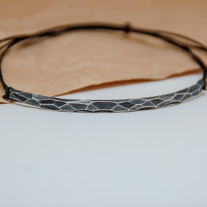Könnte beinhalten: Ein silbernes Armband mit geometrischem Muster, das an einem schwarzen Band befestigt ist.