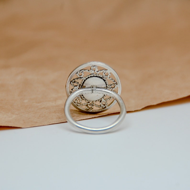 Op de afbeelding: Een zilveren ring met een filigraan ontwerp. De ring ligt op een bruine papieren achtergrond.