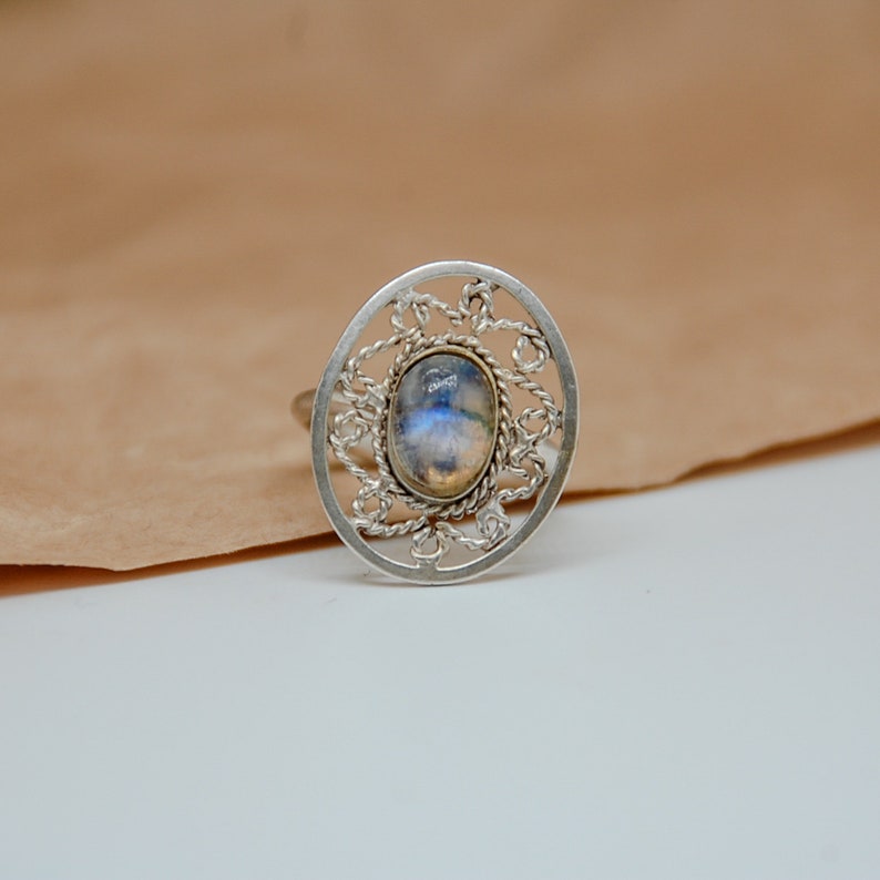Op de afbeelding: Een zilveren ring met een delicaat, ingewikkeld ontwerp met een ronde, ovale maansteen in het midden.
