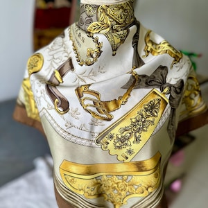 May include: A cream-coloured silk scarf with a brown border. The scarf displays ornate gold and brown decorative elements, including floral patterns, ribbons, and stylized objects. The text "PARIS" is visible on the scarf.