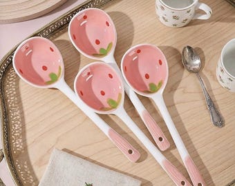 May include: Four white ceramic serving spoons with pink strawberry designs in the bowls. The spoons have pink handles and are arranged on a wooden tray. A small embroidered strawberry is on a linen napkin.