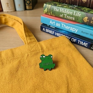 May include: A green frog-shaped enamel pin with the word "REALLY?" in white text, pinned to a mustard yellow tote bag. A stack of books is in the background, with titles including "The Hidden Life of Trees".