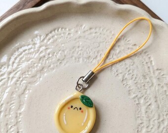 May include: A yellow lemon charm with a sad face and a green leaf. The charm is attached to a yellow cord with a silver-colored metal clasp. The charm is on a white ceramic plate.