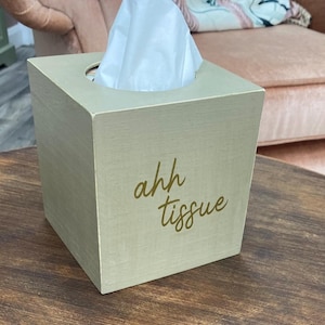 May include: A square, pale green wooden tissue box with the words "ahh tissue" in gold script. White tissues protrude from the top. The box is on a dark brown wooden table.