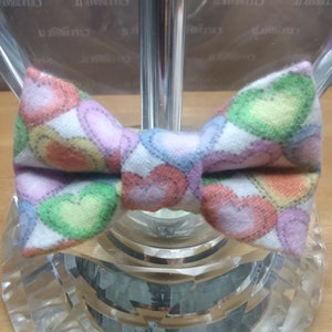 May include: A white bow tie featuring a pattern of colourful hearts. The bow tie is made from fabric and has a soft, textured feel.