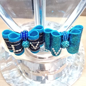 Puede incluir: Dos lazos decorativos para el cabello. Uno es turquesa con pedrería plateada, y el otro es negro y azul marino con pedrería plateada. Ambos lazos tienen un centro de cuentas azules y se exhiben sobre una base de vidrio transparente facetado.