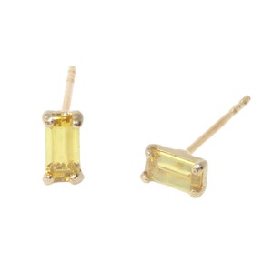 May include: A pair of gold-colored stud earrings, each featuring a rectangular, light yellow gemstone. The earrings are set in a four-prong setting, with a post for pierced ears. The gemstones have a subtle, translucent quality.