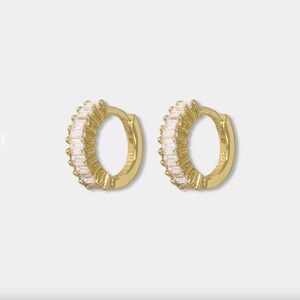 May include: A pair of gold-colored hoop earrings. Each earring is encrusted with rectangular, clear gemstones. The earrings are displayed against a plain, light background. The earrings are a jewelry item.