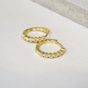 Könnte beinhalten: Ein Paar goldfarbene Creolen. Jeder Ohrring hat eine Reihe kleiner, klarer Edelsteine. Die Ohrringe werden auf einer weißen Oberfläche mit einem weichen, neutralen Hintergrund präsentiert. Die Ohrringe sind für durchbohrte Ohren konzipiert.