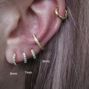 Könnte beinhalten: Nahaufnahme eines Ohrs mit mehreren goldenen Creolen. Drei kleine Reifen mit den Maßen 8 mm, 7 mm und 6 mm sind zu sehen, jeder mit kleinen, funkelnden Steinen besetzt. Ein größerer, schlichter goldener Reifen ist ebenfalls vorhanden.