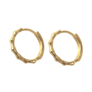 May include: A pair of gold-colored hoop earrings. Each earring has a textured, bamboo-like design around the hoop. The earrings are shown against a white background.