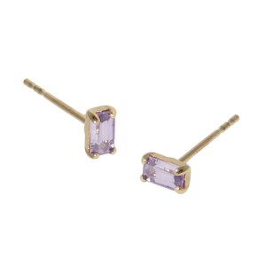 May include: A pair of gold-colored stud earrings, each featuring a rectangular, light purple gemstone. The earrings are set in a gold-colored metal and are displayed against a white background. The earrings are small and delicate.