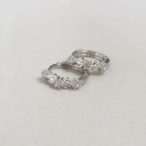 May include: Two silver hoop earrings with a floral design. Each earring features small, clear gemstones set within the flower-shaped accents. The earrings are displayed on a light-colored surface.
