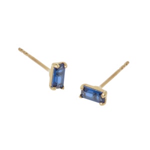 May include: A pair of gold-colored stud earrings, each featuring a rectangular, deep blue gemstone. The earrings have a simple, elegant design, with the gemstone set in a gold bezel. The posts are thin and gold-colored.