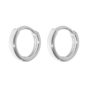 May include: A pair of small, silver-colored hoop earrings. The earrings are round with a smooth, polished surface. The hoops are designed to hug the earlobe. The earrings are displayed against a white background.