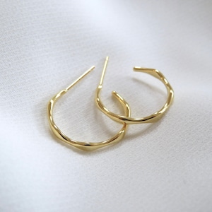May include: A pair of gold-colored hoop earrings with a textured, irregular surface. The earrings have a post closure and a slightly flattened, organic shape. The background is a soft, white fabric.