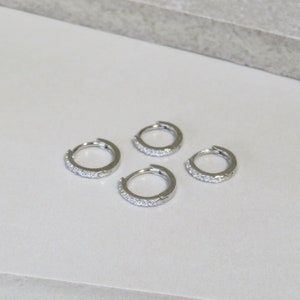 May include: Four small, silver-colored hoop earrings. Each earring is adorned with a row of small, clear gemstones. The earrings are arranged on a light-colored surface, showcasing their simple yet elegant design.