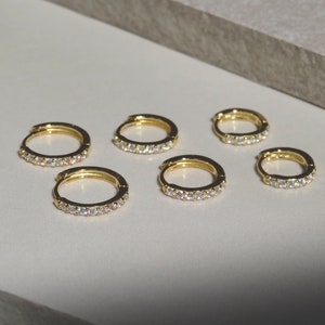 May include: Six gold-colored hoop earrings with small, clear gemstones set along the outer edge. The earrings are arranged on a light-colored surface, with a neutral background.