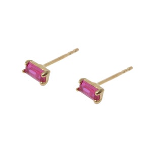 May include: A pair of gold-colored stud earrings. Each earring features a rectangular, bright pink gemstone set in a gold-colored bezel. The earrings are small and delicate, with a simple, elegant design.