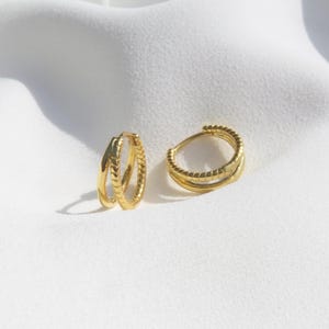 May include: Two gold-colored hoop earrings. Each earring has a double-hoop design with a textured, ridged pattern. The earrings are displayed on a white, textured surface, creating a contrast that highlights their metallic sheen. The earrings are a jewelry item.