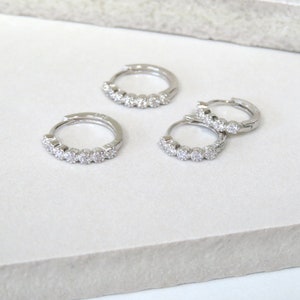May include: Four silver hoop earrings, each adorned with a row of small, sparkling clear gemstones. The earrings are arranged on a white surface, showcasing their reflective qualities and delicate design. The earrings are a classic jewelry item.