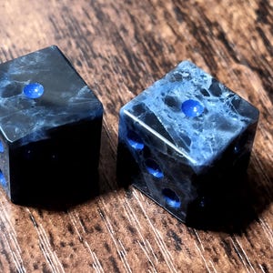 Könnte beinhalten: Zwei Würfel mit einem marmorierten schwarz-blauen Design. Jeder Würfel hat blaue Punkte, die die Zahlen anzeigen. Die Würfel sind vor einem Holzgrund platziert. Diese Würfel eignen sich für Tischspiele.