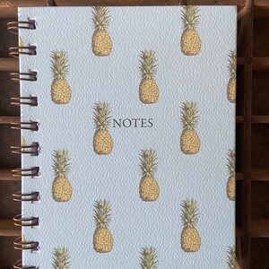 Puede incluir: Una libreta de espiral azul claro con un patrón de piña y la palabra "NOTES" impresa en negro en la portada.