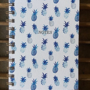 Puede incluir: Un cuaderno con espiral con una cubierta blanca con un patrón repetido de piñas azules en acuarela. La palabra "NOTES" está impresa en negro. El cuaderno tiene una encuadernación en espiral dorada.