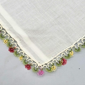 May include: A white square textile with a visible woven texture, featuring a decorative needle lace border. The edging consists of small, repeating loops in cream, accented with tiny floral motifs in shades of olive green, yellow, red, and pink.
