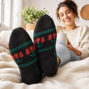 Puede incluir: Un par de calcetines de punto negro con un patrón repetitivo de flores rojas con tallos y hojas verdes. Los calcetines tienen una apariencia tejida y texturizada con una banda horizontal de detalles geométricos verdes por encima y por debajo del diseño floral.