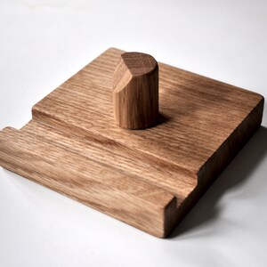 May include: A wooden phone stand with a square base and a rounded top. The stand is made of light brown wood and has a groove for holding a phone.