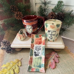 May include: A red ceramic mug with the text "be JOYFUL ALWAYS", a cream-colored ceramic cup with the text "HE RESTORES MY SOUL", and a rectangular ceramic tray with the text "so now FAITH HOPE and LOVE abide, these three; but the greatest of these is LOVE".